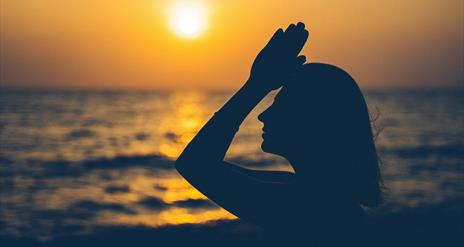 a person raises joined hands to their forehead with the sunset and the sea in the background