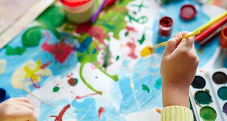 Child's hands paint across a messy canvas, surrounded by various art supplies.