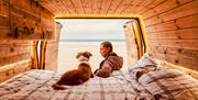 A woman and her dog lying on the bed of a campervan, looking out of the open back doors at the beach