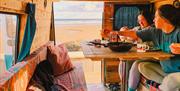 Two friends having breakfast at the table inside a campervan, with the sea through the open door