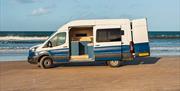 A blue and white Karen & Co. campervan by the sea with its side door open, showing the fitted kitchen inside