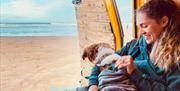 A woman cuddling her dog in the open doorway of a campervan, with the beach behind