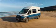 A blue and white Karen & Co. campervan parked on a wide sandy beach below green cliffs