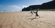 Image of beach with cliffs and sea in the background. The iconic Mussenden Temple is visible in the distance. In the foreground, 'welcome' has been ca