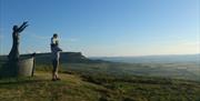 Person standing beside Manannan Mac Lir statue looking at fields and blue sky.