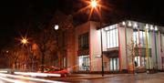 A photo of the building taken at night. Street lights and cars with the building lit up in the background.