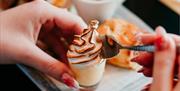 a person's hand uses a spoon to lift a small meringue-style dessert from its container which sits on a plate with sandwiches, a sausage roll and a sma
