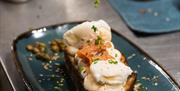 a persons hand sprinkles a herb garnish over poached eggs and salmon on sourdough bread