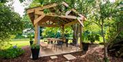 The Garden Cookhouse gazebo at The Willows featuring timber framing, outdoor dining table, benches, and surrounding trees.