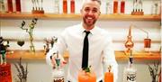 Expert Cocktail Maker Ashley stands in shirt and black tie smiling in front of a beautiful bar consisting of a white wall, 3 wooden shelves topped wit