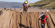 Two riders jumping a sand hill with the beach and people in the background