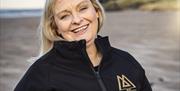 Claire O'Kane of Mussenden Sea Salt stands smiling beneath Mussenden Temple, wearing a black zip up fleece with her company logo in gold.