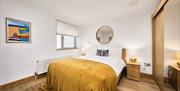 Photograph of double bedroom with mustard velour throw, 2 bedside cabinets and 2 bedside lights. Wall art of beach scene on the wall.