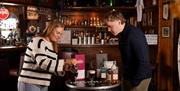 a woman pours cream in to an Irish Coffee sitting on a barrel table at a bar, while a man watches on