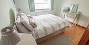 Double bed with neutral striped bedding in a bright bedroom with wooden floor, window, and dressing table.