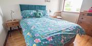 Double bed with teal floral bedding in a bright bedroom with wooden floor and window.
