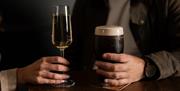 a woman's hand holds a glass of prosecco while a man's hand holds a pint of black stout