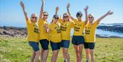 Five women in yellow Giant Steps t-shirts standing on a grassy coastal path with the sea behind them, raising their arms and smiling in celebration.