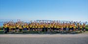 A large group of Giant Steps participants wearing yellow t-shirts gathered outdoors by the coast, raising their hands together in a wide group photo.