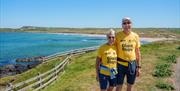 Two participants in yellow Giant Steps t-shirts standing on a coastal path with a wooden fence and blue sea behind them, smiling at the camera.