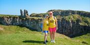 Two participants in yellow Giant Steps t-shirts standing on a grassy cliff with rugged coastal rocks behind them, smiling during the event.