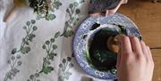 a persons hands dips a wooden stamp in to a bowl of green dye, which sits a top a piece of linen with printed foliage