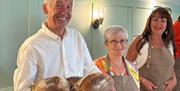 A man stands smiling holding loaves of fresh sourdough bread. Two women in aprons stand behind him and to the right.