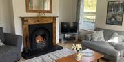 Bright, comfortable living room with a lit fireplace, wooden mantel and mirror, grey armchairs and sofa, a coffee table with flowers, and a window bes