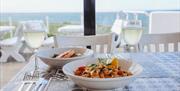 two bowls of food and two glasses of wine sit on a mosaic style table with a view out to sea