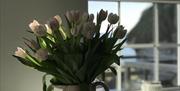 A vase of tulips on dining table with window in background looking out towards the deck, beach and sea.