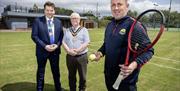 3 men stand for a photo on a tennis court, one is holding a tennis racket and tennis ball