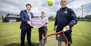 2 men hold a sign saying 'Event Fund' while another bounces a tennis ball on a racket