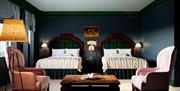 Spacious hotel room with two double beds featuring tartan headboards, soft cream bedding, and classic wood furniture. The room includes pink armchairs