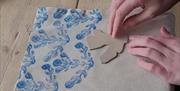 a person's hands used a block to print a blue floral design on to a piece of linen