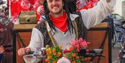 a man dressed as a pirate cycles on a bike adorned with flowers while bubbles float from a box he is towing behind him