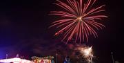 Image shows fireworks lighting up the night sky with the crowd and fairground amusements below