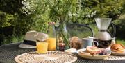 Table on hut deck laid with breakfast items - orange juice pourded, bread sliced with jam, croissants, compote, and freshly brewed coffee.
