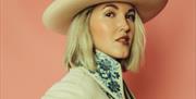 Singer Ashley Campbell is pictured wearing a large embroidered collared shirt and a cream hat against a peach coloured background