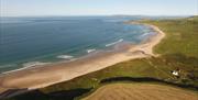 The photo is of White Park Bay beach taken as an aerial photo. There are green areas to the right of the beach and a small cottage.