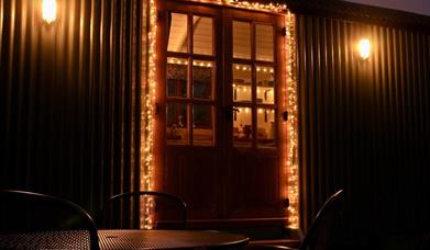 exterior of Peatlands self-catering cabin at night. Fairy lights surround the main door with two sconces lit on either side.