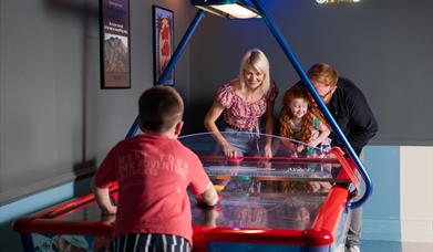 a family are gathered around an air hockey table at the arcade at Roe Valley Resort