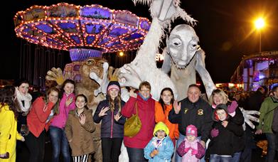 Image shows a group of people standing in front of scary Halloween characters, with a Fairground in the background 