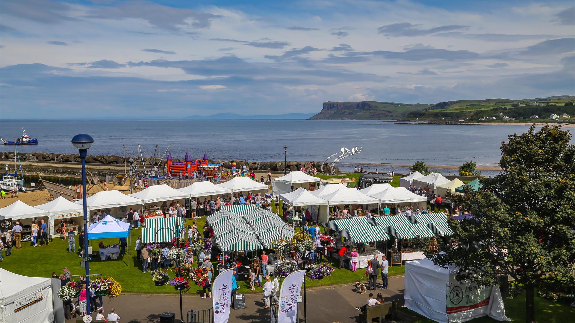 Overhead Image showing coastline at Ballycastle, sea in background.  Foreground shows green and white striped tents, people browsing round the markets