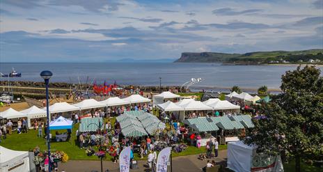 Overhead Image showing coastline at Ballycastle, sea in background.  Foreground shows green and white striped tents, people browsing round the markets