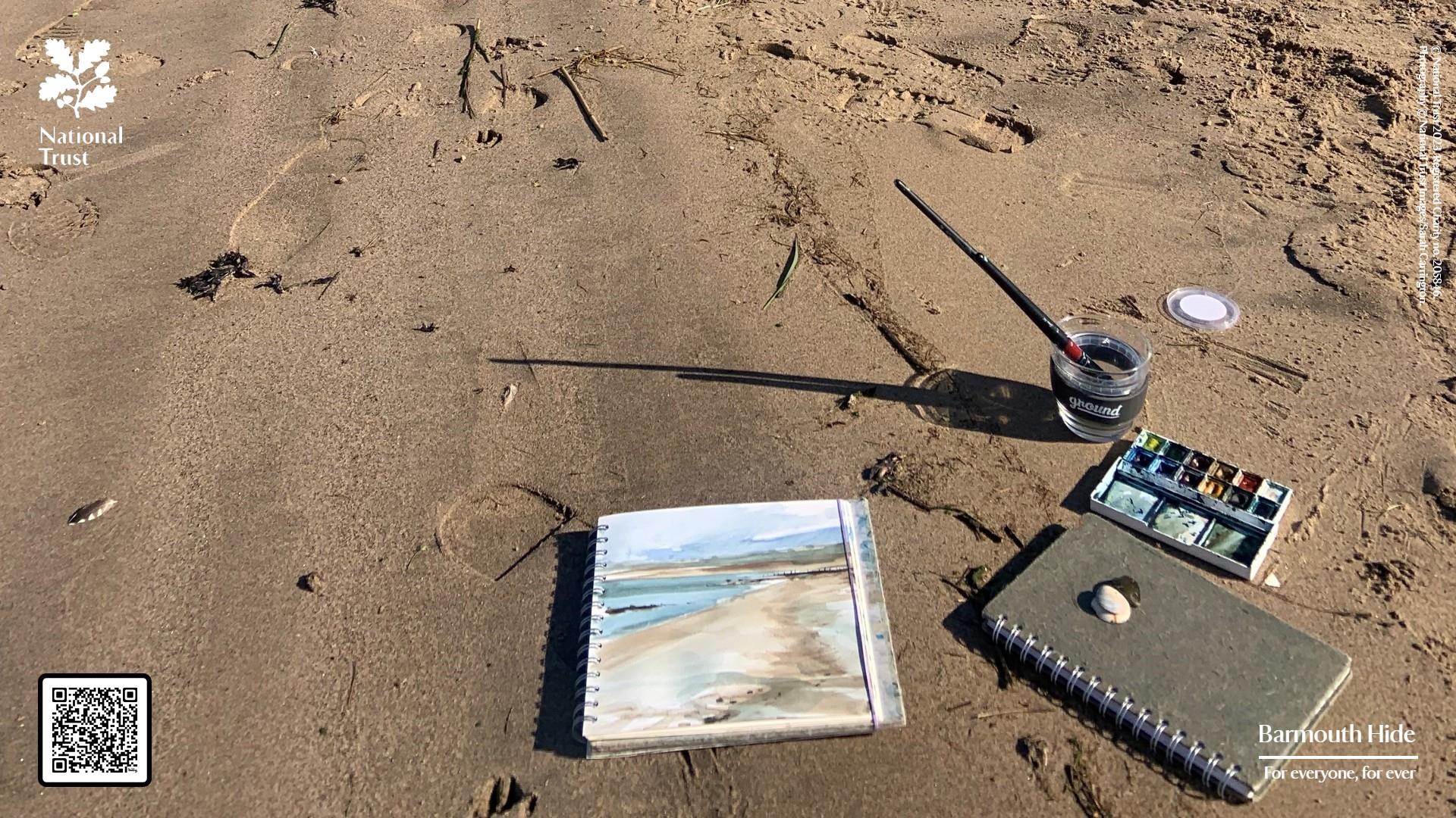A notepad with a watercolours painted on the page lies on the beach.
