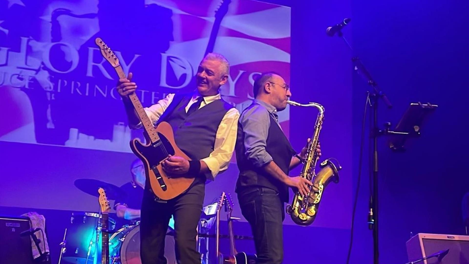 musicians from the Glory Days Bruce Springsteen Tribute band perform on stage. One is strumming a guitar while the other plays a saxophone, and a drum musicians from the Glory Days Bruce Springsteen Tribute band perform on stage. One is strumming a guitar while the other plays a saxophone, and a drum