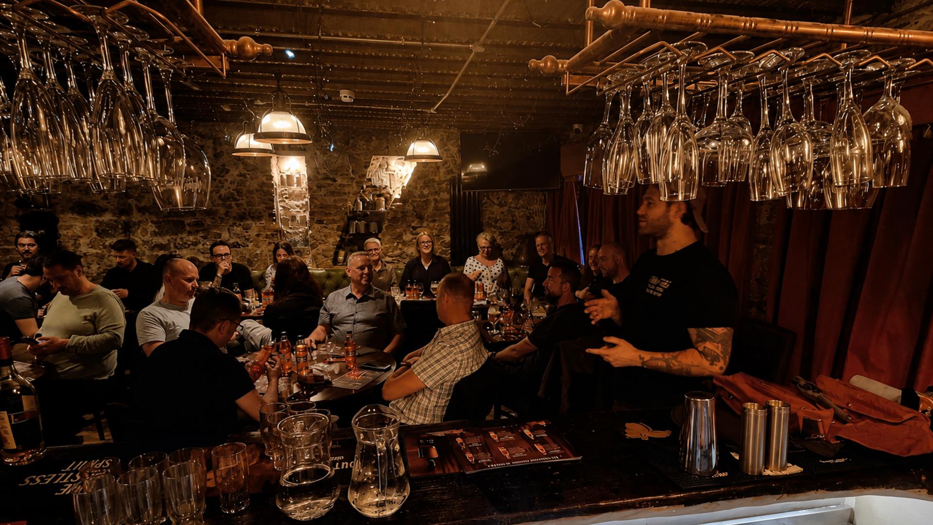 a large group of people sit around tables in a dimly lit bar listening to a man speaking who is standing a large group of people sit around tables in a dimly lit bar listening to a man speaking who is standing