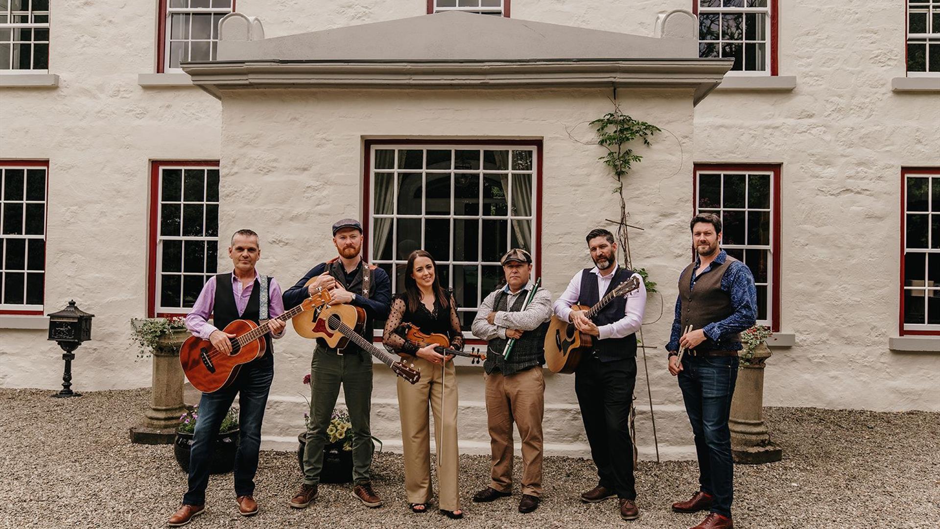 Image shows the six members of the band standing in front of a white house holding instruments