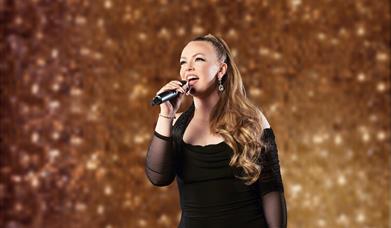 Image shows a woman wearing a black dress, singing into a microphone in her hand and standing in front of a gold glitter wall