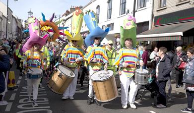 Carnival parade makes its way down the street with performers dressed in bright yellow costumes wearing tall coloured hats